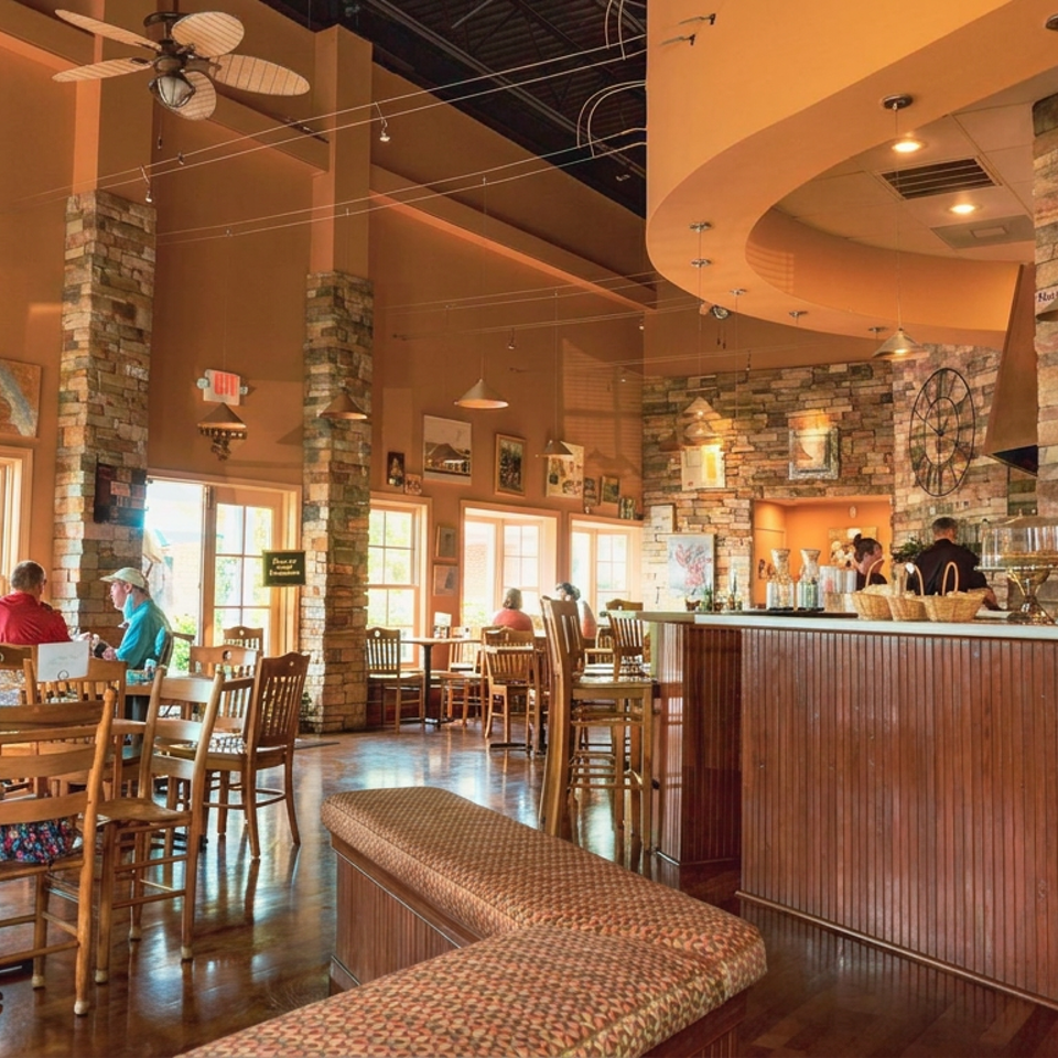 Warmly lit café interior with stone columns, wooden tables, seated customers, and a long counter displaying pastries and beverages.